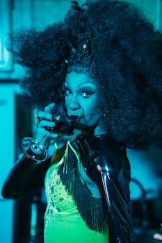 Stylish African American transvestite in curly wig drinking wine from glass in bar while looking at camera